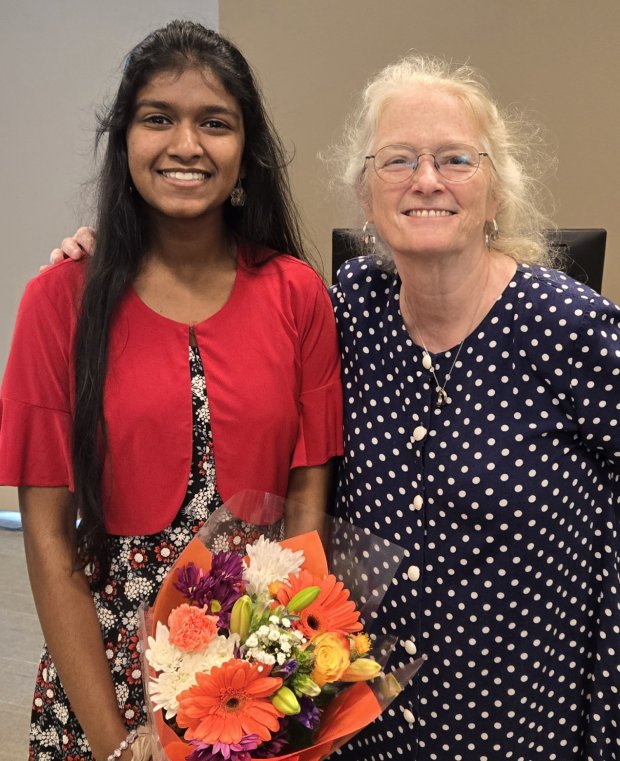 A young woman with long dark hair wearing a red shrug and holding a bouquet of flowers smiles for a picture with a woman with blond hair, glasses and a polka dot top.