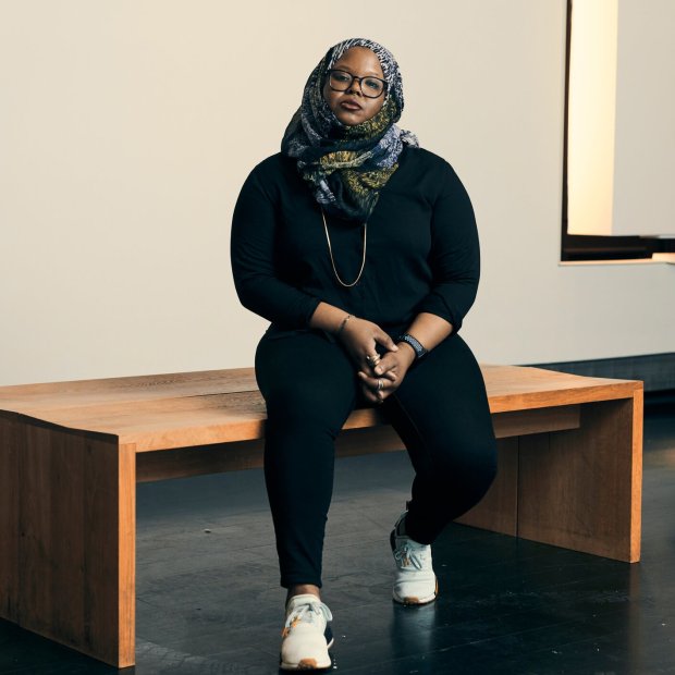 Person in a headscarf and glasses sitting on a bench in a gallery space