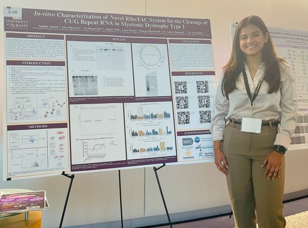 A young woman with long dark hair wearing a lanyard and cream silk top stands next to a poster titled “In vitro characterization of novel riboTAC system for the cleavage of CUG repeat RNA in myotonic dystrophy type 1."