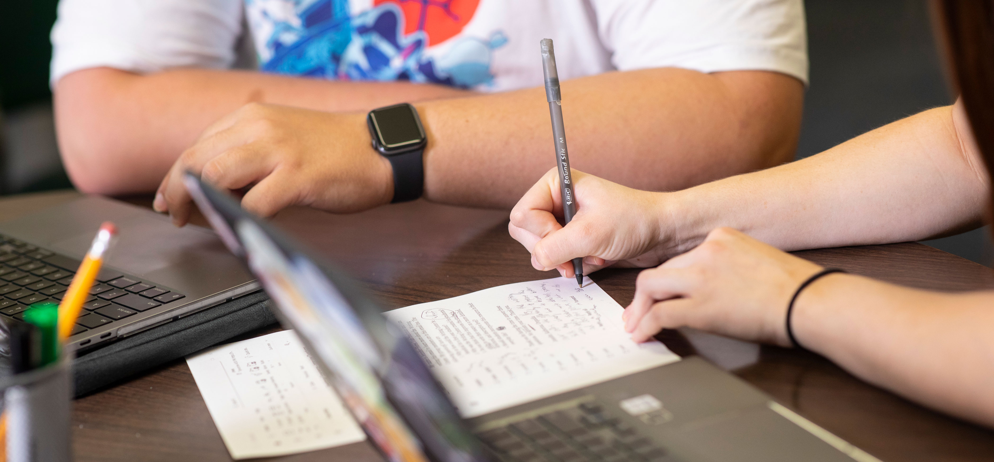 A close-up look at two people's hands as they work to edit an essay together. Two laptops are open on the table as they work with pen and paper.