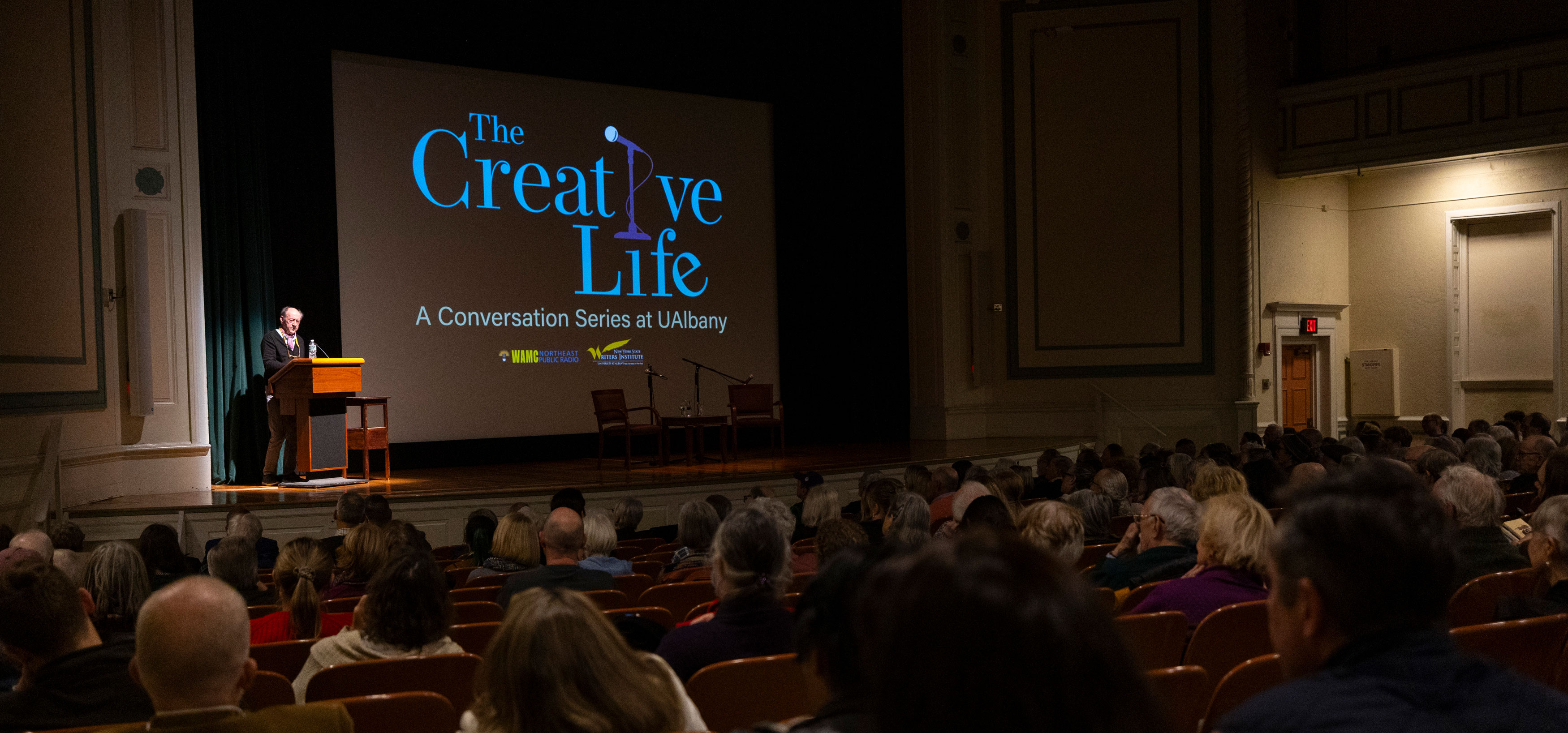 A man stands at a podium and speaks to an auditorium full of people, with the on-stage projector showing a slide with the words, "The Creative Life, A Conversation Series at UAlbany."